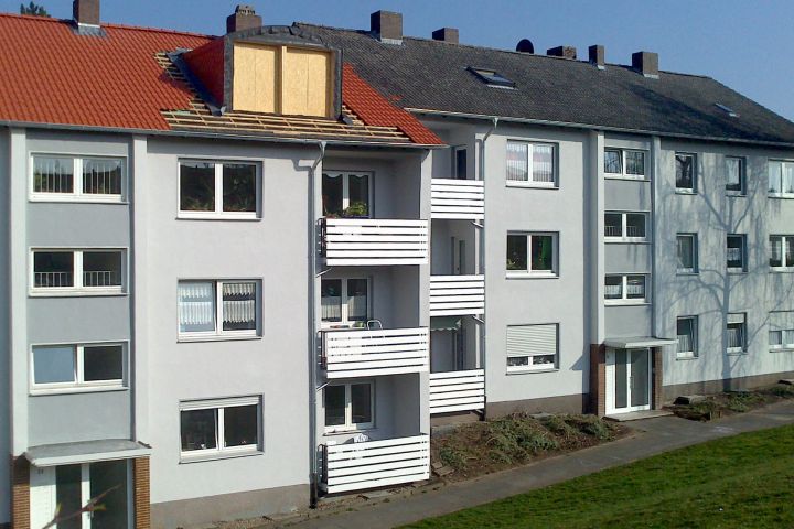 Mehrfamilienhaus mit grauer Fassade, weißen Balkonen und einem roten Dach in Renovierung.
