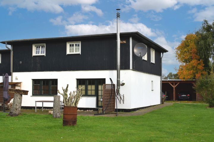 Zweistöckiges Haus mit schwarzem Obergeschoss, Garten und Carport im Hintergrund.