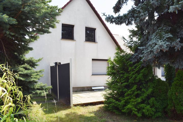 Weißes Haus mit Satteldach, umgeben von Bäumen und Sträuchern, Holzterrasse vorne.