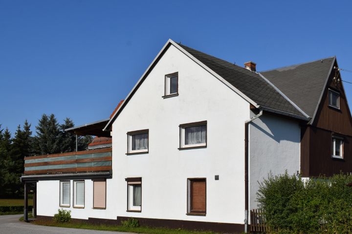 Weißes Haus mit Satteldach, braunen Fensterläden, Balkon und blauen Himmel im Hintergrund.