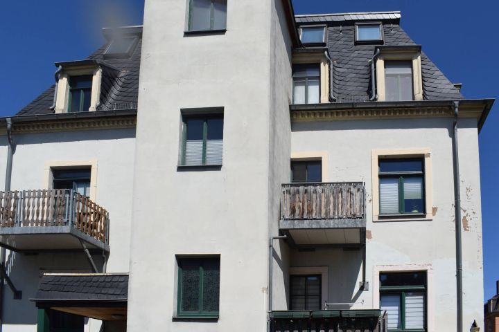 Mehrstöckiges Gebäude mit grauem Dach, Balkonen und grünen Fensterrahmen.