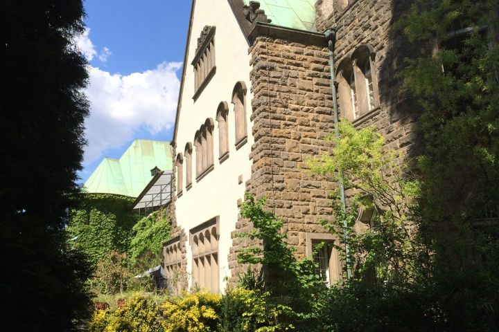 Steinbau mit grünen Dächern, umgeben von Bäumen und Pflanzen bei sonnigem Wetter.