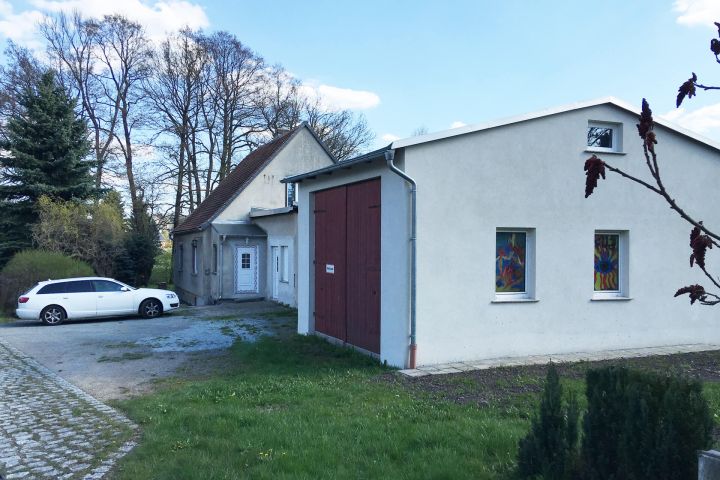 Ein weißes Auto steht vor zwei Gebäuden, umgeben von Bäumen und grünem Rasen.