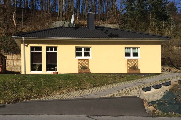 Gelbes Haus mit flachem Dach und zwei großen Fenstern, umgeben von Bäumen.