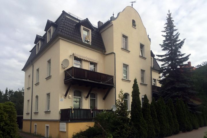 Mehrstöckiges, beige Gebäude mit Dachgauben, Balkon und umgeben von Bäumen.